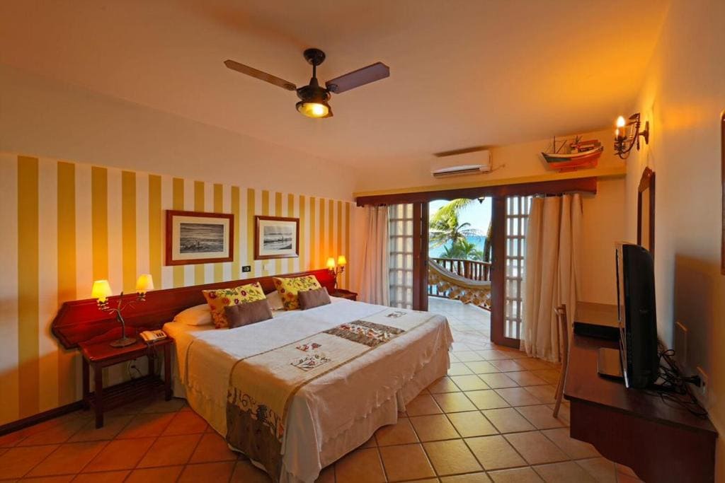 Intérieur d'une chambre d'hôtes élégante au Manary Praia Hotel Natal avec un grand lit, une décoration aux tons jaunes et bois, et un balcon vue mer avec hamac.
