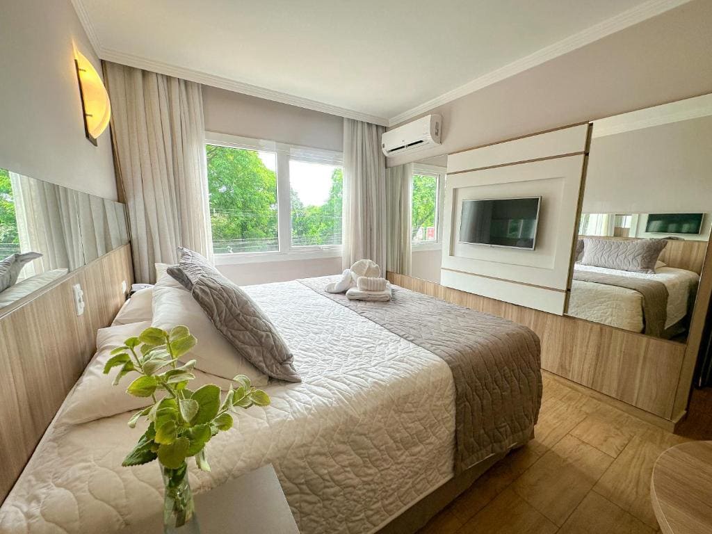 Intérieur d'une suite moderne au Nadai Confort Hotel e Spa avec un grand lit blanc et gris, une télévision murale sur panneau de bois et une vue sur la nature.