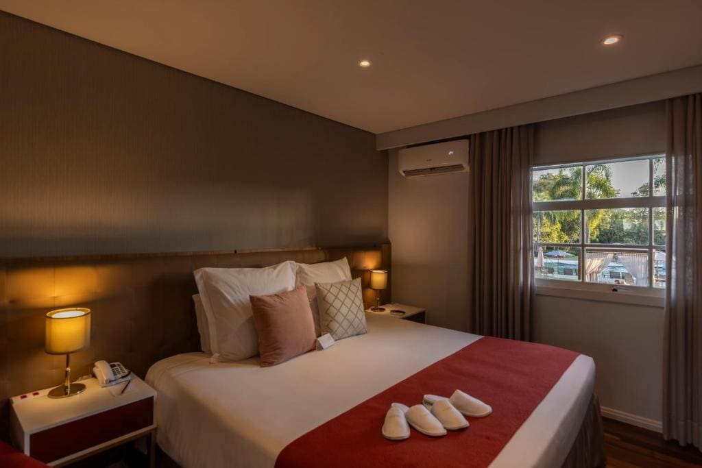 Chambre d'hôtel luxueuse à l'Hotel Sanma Iguaçu, avec grand lit, éclairage tamisé, murs marrons et vue sur une piscine et des jardins extérieurs.
