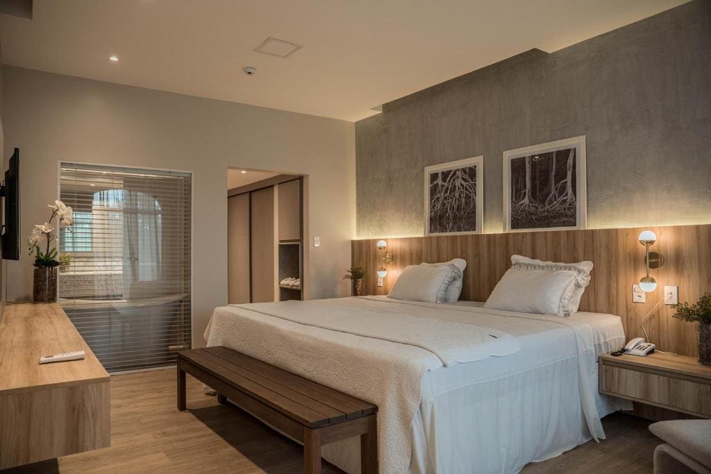 Photo d'une chambre moderne de l'hôtel Blue Tree de São Luís avec un grand lit double fait de blanc, une tête de lit en bois sous un éclairage doux, deux tableaux au mur, un banc en bois au pied du lit et une verrière donnant sur la salle de bain.