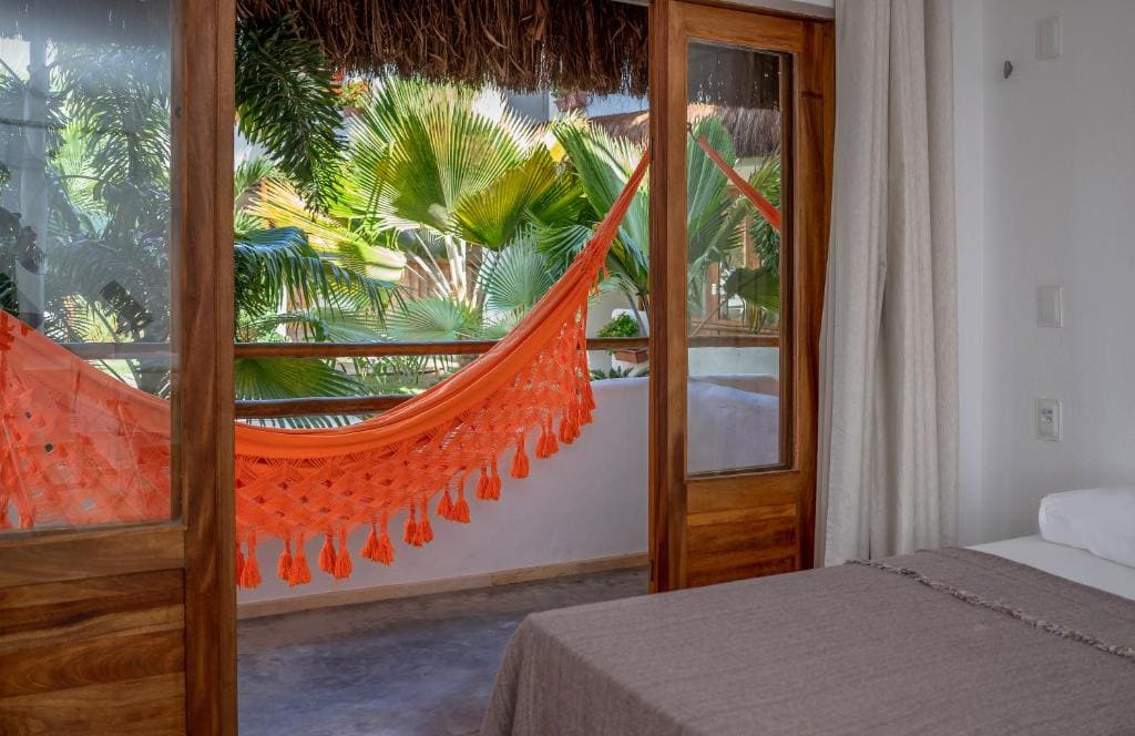 Intérieur d'une chambre d'hôtel à Jericoacoara ouvrant sur un balcon avec un hamac orange et des palmiers.