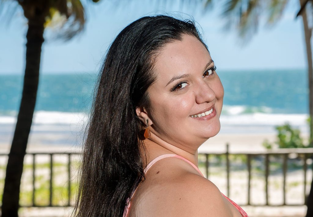 Portrait de Gloria, responsable de production pour l'agence Brazil Selection, souriante en robe rose avec un fond de plage tropicale et de palmiers.