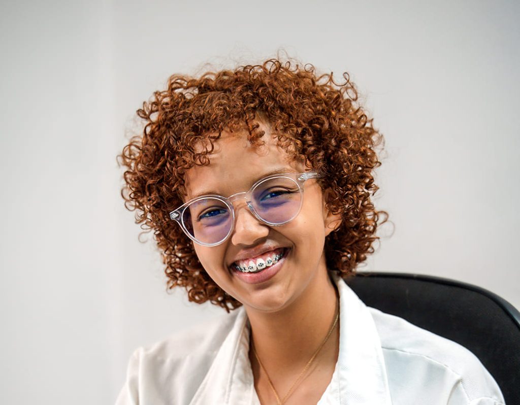 Portrait en gros plan de Gabrielle, chargée de la coordination et du contrôle qualité pour l'agence Brazil Selection, souriante avec des cheveux bouclés châtains, des lunettes transparentes et une chemise blanche, sur fond clair.