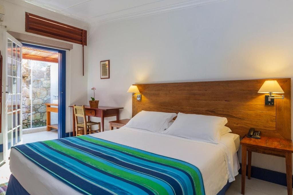 Intérieur d'une chambre d'hôtel avec un grand lit central. Le lit a des draps blancs et une jetée de lit aux rayures bleues et vertes. Derrière le lit se trouve une grande tête de bois et deux appliques murales. À droite, une table de nuit avec une lampe. À gauche, un bureau et une chaise près d'une porte en verre donnant sur un patio. Le mur est blanc. L'éclairage est chaleureux.