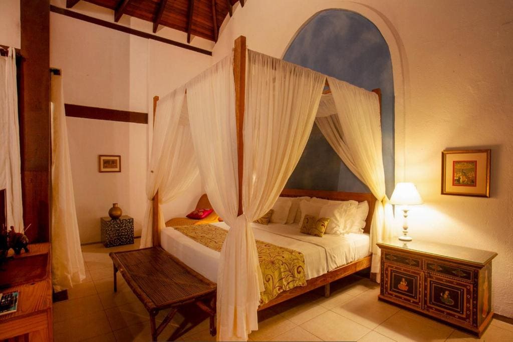 Intérieur d'un bungalow de luxe. Un grand lit à baldaquin avec des voilages blancs est au centre, dressé avec des draps blancs et un jeté de lit doré. Le mur du fond est blanc avec une alcôve bleue. À droite, un meuble décoré sert de table de nuit avec une lampe allumée. Un banc en bois est au pied du lit. Le plafond est en bois avec des poutres apparentes. Le sol est carrelé. L'éclairage est chaleureux et tamisé.