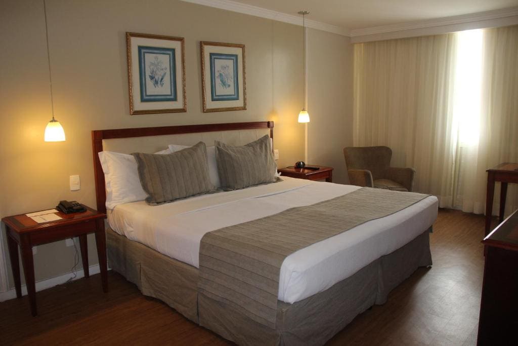 Intérieur d'une chambre d'hôtel avec un grand lit central fait avec des draps blancs et des jetés taupe. Des tables de chevet en bois et des lampes suspendues encadrent le lit. Au fond, deux cadres avec des motifs botaniques sont accrochés au mur. Sur la droite, un fauteuil confortable est près d'une fenêtre avec des rideaux clairs. Le sol est en parquet bois. L'ambiance est chaleureuse et classique.