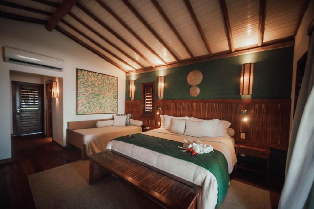 Intérieur d'une chambre d'hôtel avec un grand lit central et un canapé-lit sur la gauche. Le lit a une jetée de lit verte et des coussins blancs. Le mur derrière le lit est vert avec des décorations murales en bois. Le plafond est en bois avec des poutres apparentes. Un grand tableau coloré est accroché au-dessus du canapé. Une grande fenêtre sur la droite donne sur la mer. Le sol est en bois avec un tapis.