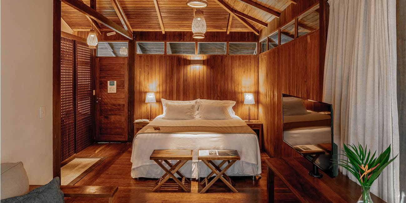 Intérieur d'un chalet tout en bois, avec un grand lit central fait avec des draps blancs et des jetés de lit. Deux tables de chevet et des lampes de table encadrent le lit. Des suspensions tressées pendent du plafond en bois apparent. Deux petits tabourets en bois sont devant le lit. Sur la droite, un téléviseur est sur une console avec une plante. Une porte en bois et un claustra en bois décorent la pièce. Le sol est en bois. L'éclairage est chaleureux et tamisé.