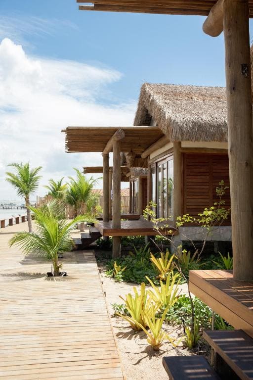 Vue extérieure de bungalows en bois avec des toits en paille au K08 Bangalôs à Galinhos. Une passerelle en bois longe les hébergements sur pilotis, entourés de végétation tropicale et de sable. Le ciel est bleu avec quelques nuages.