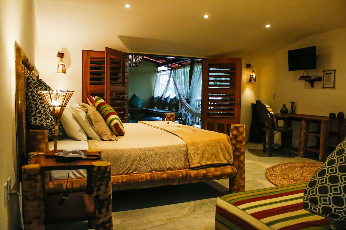 Intérieur d'une chambre d'hôtel rustique avec un grand lit central. La tête de lit et le cadre du lit sont faits de troncs d'arbres. Les draps sont blancs avec des coussins colorés. De chaque côté du lit se trouvent des tables de nuit en bois et des lampes artisanales. Au fond, de grandes portes en bois s'ouvrent sur une terrasse couverte avec un hamac et des sièges. Un bureau en bois et une télévision sont sur la droite. Le sol est en béton. L'éclairage est tamisé.