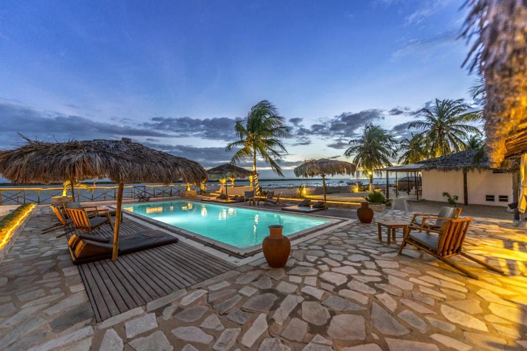 Terrasse en pierre d'une pousada au crépuscule, avec une piscine éclairée de l'intérieur, des chaises longues sous des parasols en paille, des palmiers et un ciel de fin de soirée.