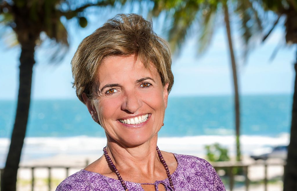 Portrait en gros plan d'Aude, conseillère voyages pour l'agence Brazil Selection, souriante avec un collier violet sur fond de plage tropicale et de palmiers.