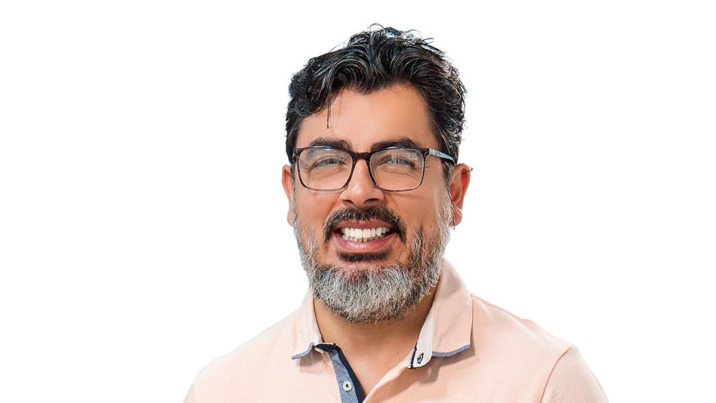 Portrait de Fernando, directeur administratif et financier pour l'agence Brazil Selection, souriant avec des lunettes, une barbe grisonnante et un polo rose clair, sur fond blanc.