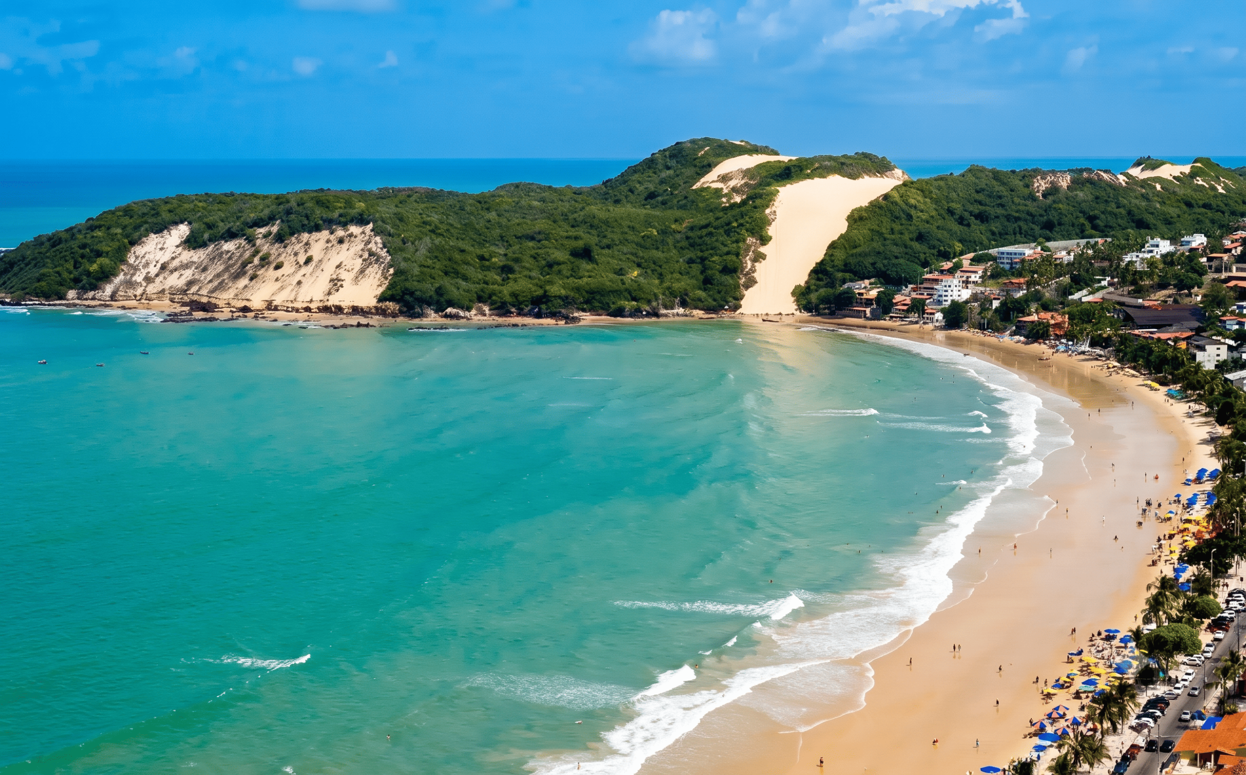 Photo aérienne d'une plage en forme de croissant bordée d'un côté par une mer turquoise et de l'autre par une ville balnéaire. Une grande dune couverte de végétation se dresse à une extrémité de la plage. La plage est remplie de gens et de parasols colorés.