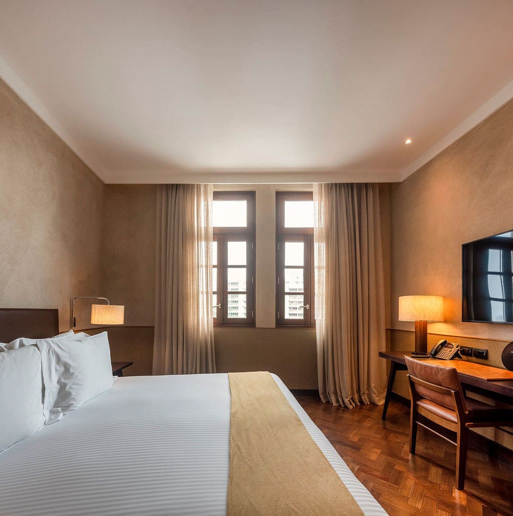 Intérieur d'une chambre d'hôtel de catégorie supérieure. Un grand lit central avec des draps blancs est sur la gauche. Au fond, deux fenêtres avec des rideaux donnent sur l'extérieur. Sur la droite, un bureau en bois et une chaise en cuir sont installés sous une lampe de table, à côté d'une télévision murale. Le mur est texturé et le sol est en parquet à chevrons. L'éclairage est chaleureux et tamisé.