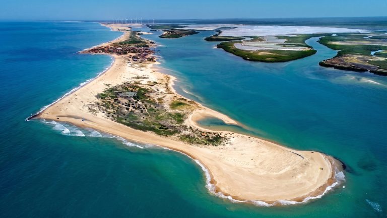 Photo aérienne d'une fine péninsule de sable doré s'avançant dans une mer turquoise et limpide. La langue de terre sépare l'océan agité à gauche d'un estuaire calme aux teintes vertes et bleues à droite. Au loin, on aperçoit un petit village de pêcheurs et un parc d'éoliennes sur la côte sous un ciel bleu sans nuage.