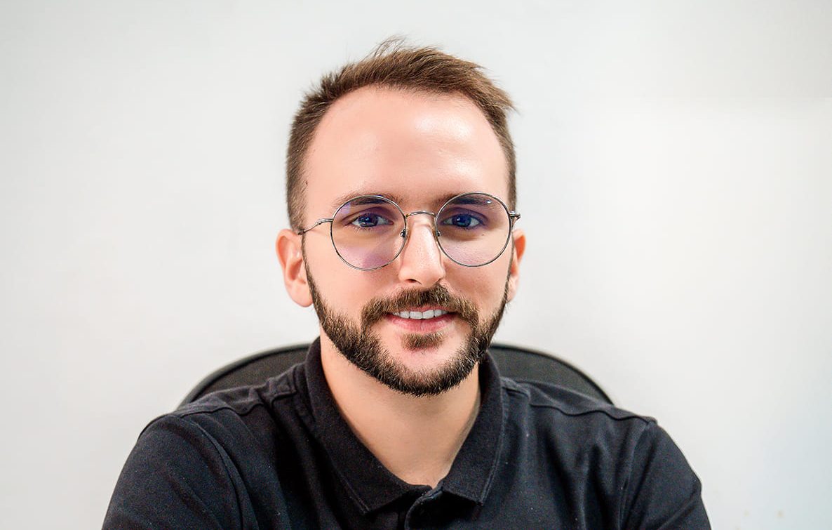 Portrait de Jonathan, conseiller en vente et développement chez Brazil Selection, portant des lunettes rondes et un polo noir sur fond clair.