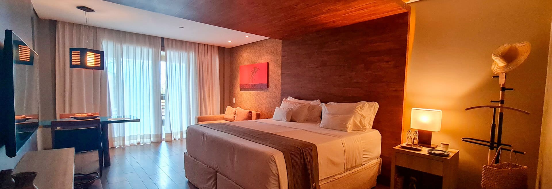 Intérieur d'une suite d'hôtel avec un grand lit central. Le mur derrière le lit est en bois avec une œuvre d'art rouge. À droite, une table de nuit avec une lampe et un portant à vêtements original avec un chapeau et un panier. À gauche, un canapé et une table basse près d'une fenêtre. Le sol est en parquet. L'éclairage est chaleureux et tamisé.