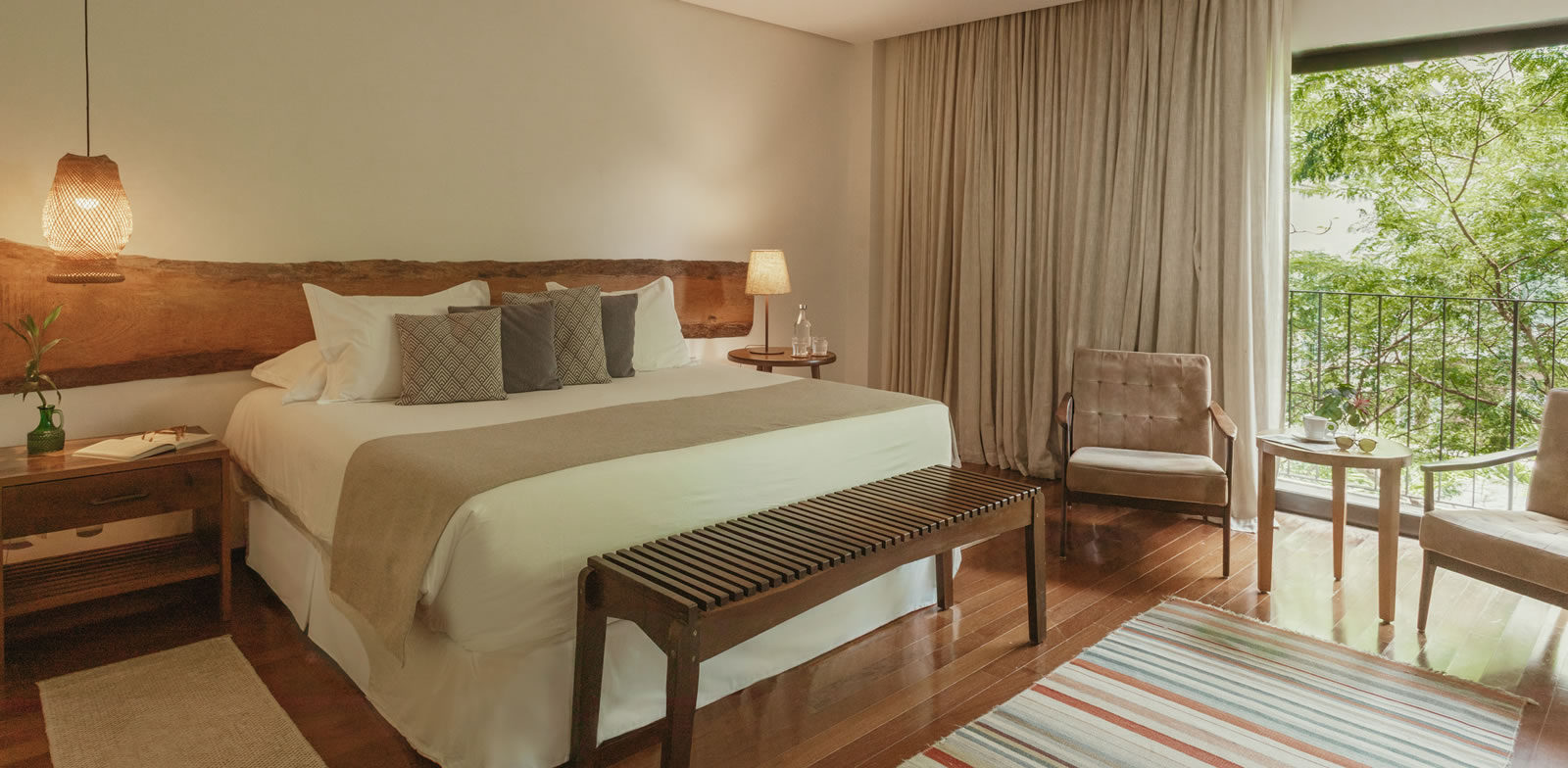 Intérieur d'une chambre d'hôtel avec un grand lit central et des tables de chevet en bois. Un mur est en grande partie constitué de rideaux qui s'ouvrent sur un balcon avec vue sur les arbres. Un coin salon avec des fauteuils est présent près de la fenêtre. Le style est chaleureux avec des matériaux naturels comme le bois et le tissage. Le sol est en bois et couvert d'un tapis.