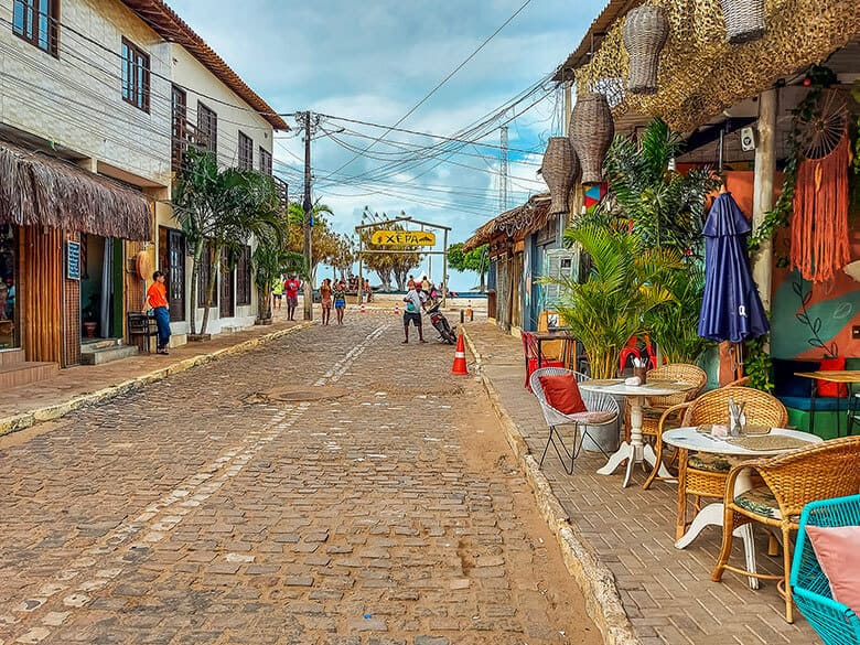 Photographie d'une rue en pavés à São Miguel do Gostoso, au Brésil, la Rua da Xêpa. La rue est bordée de bâtiments colorés, certains avec des toits de chaume, abritant des commerces et des restaurants. Au premier plan, une terrasse de café est installée avec des tables et des chaises. Des gens marchent sur la rue en arrière-plan, sous un ciel nuageux. Un panneau en bois indique "XÊPA".