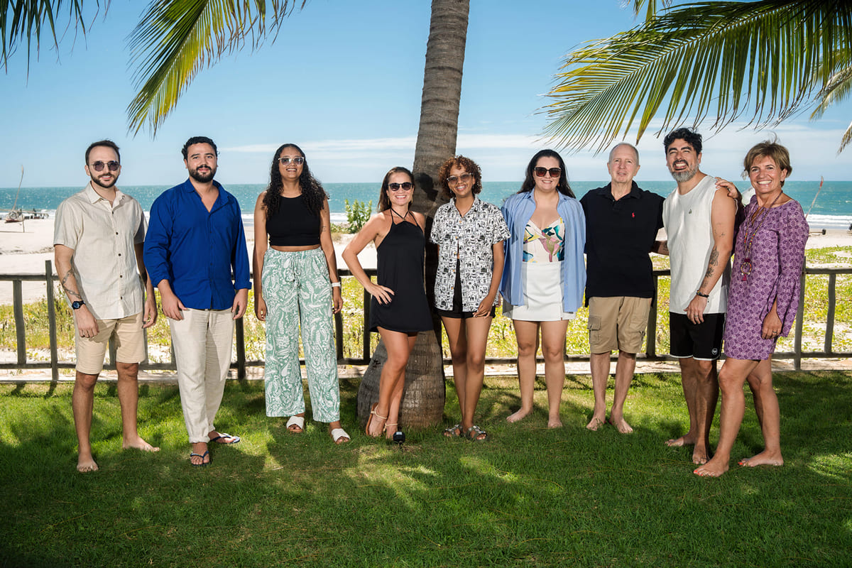 Les neuf membres de l'équipe Brazil Selection alignés et souriants sur de la pelouse devant un cocotier, avec la plage et la mer en arrière-plan par beau temps.