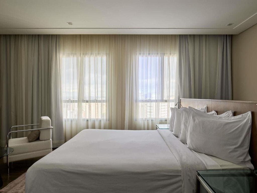 Vue d'une chambre d'hôtel moderne au Mercure Belo Horizonte Lourdes avec un grand lit aux draps blancs, des fenêtres illuminées par la lumière naturelle avec rideaux voilés, un fauteuil design et une table de chevet en verre.