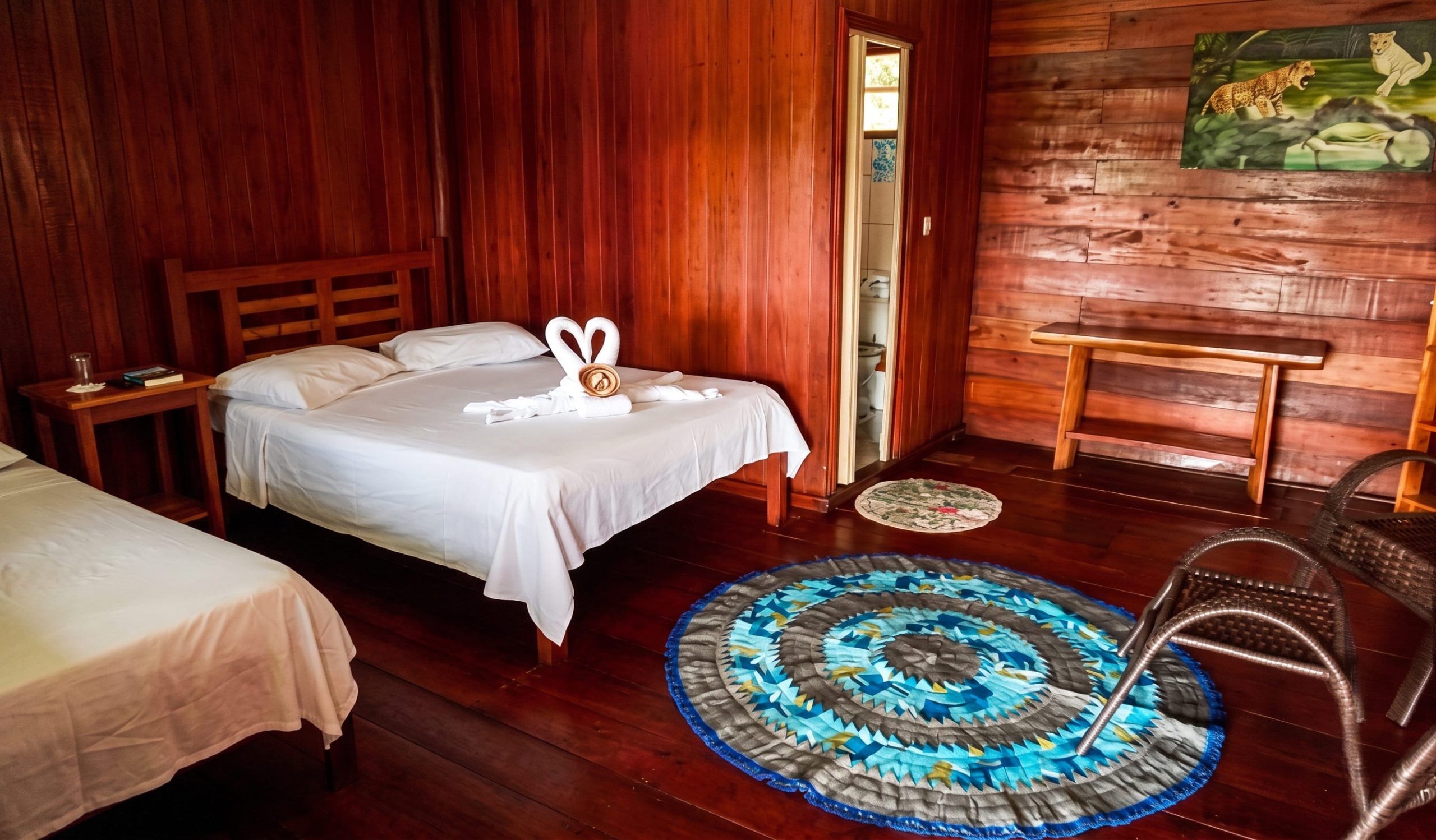 Vue intérieure grand angle d'une chambre d'hôtel au Tupana Lodge, en Amazonie. La pièce est entièrement lambrissée de bois rouge et comporte un parquet en bois sombre. Un grand lit double avec des draps blancs et des serviettes en forme de cygne est au centre, flanqué d'une table de nuit. À gauche, un lit simple est visible. Au centre du sol se trouve un tapis circulaire tissé aux motifs concentriques bleus et gris. À droite, deux chaises en rotin tissé marron font face au lit. Sur le mur du fond, un bureau en bois massif est placé sous un tableau représentant un jaguar tacheté et son petit dans la jungle. Une porte ouverte révèle une petite salle de bain. La lumière naturelle pénètre par une fenêtre sur le côté gauche.